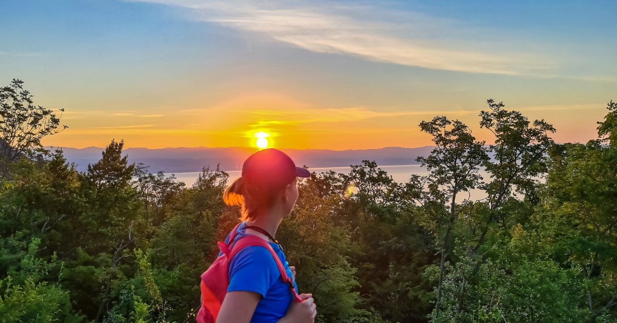 A woman with a backpack pauses on a forest overlook, watching a colorful sunset over distant mountains and water.