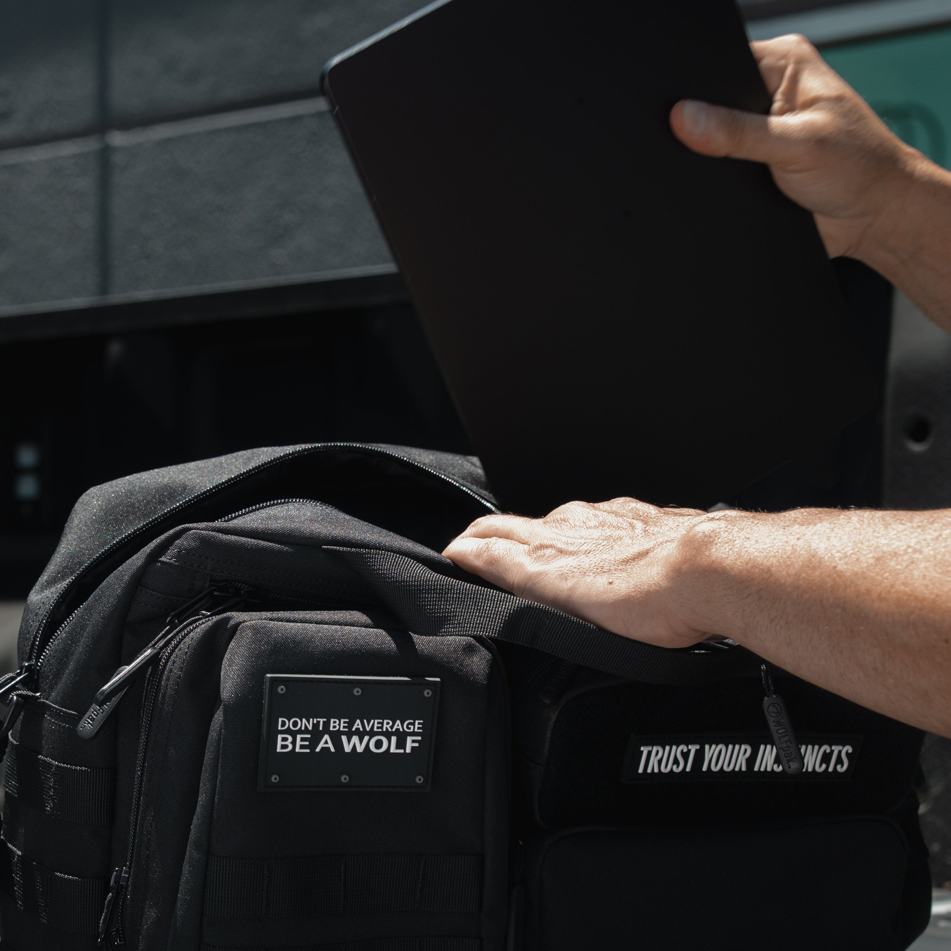 A black tactical-style laptop bag with a front patch reading 'DIGITAL WOLF IS A WOLF' and white text on the side, shown outdoors on a truck bed with a person inserting a black laptop into the main compartment.