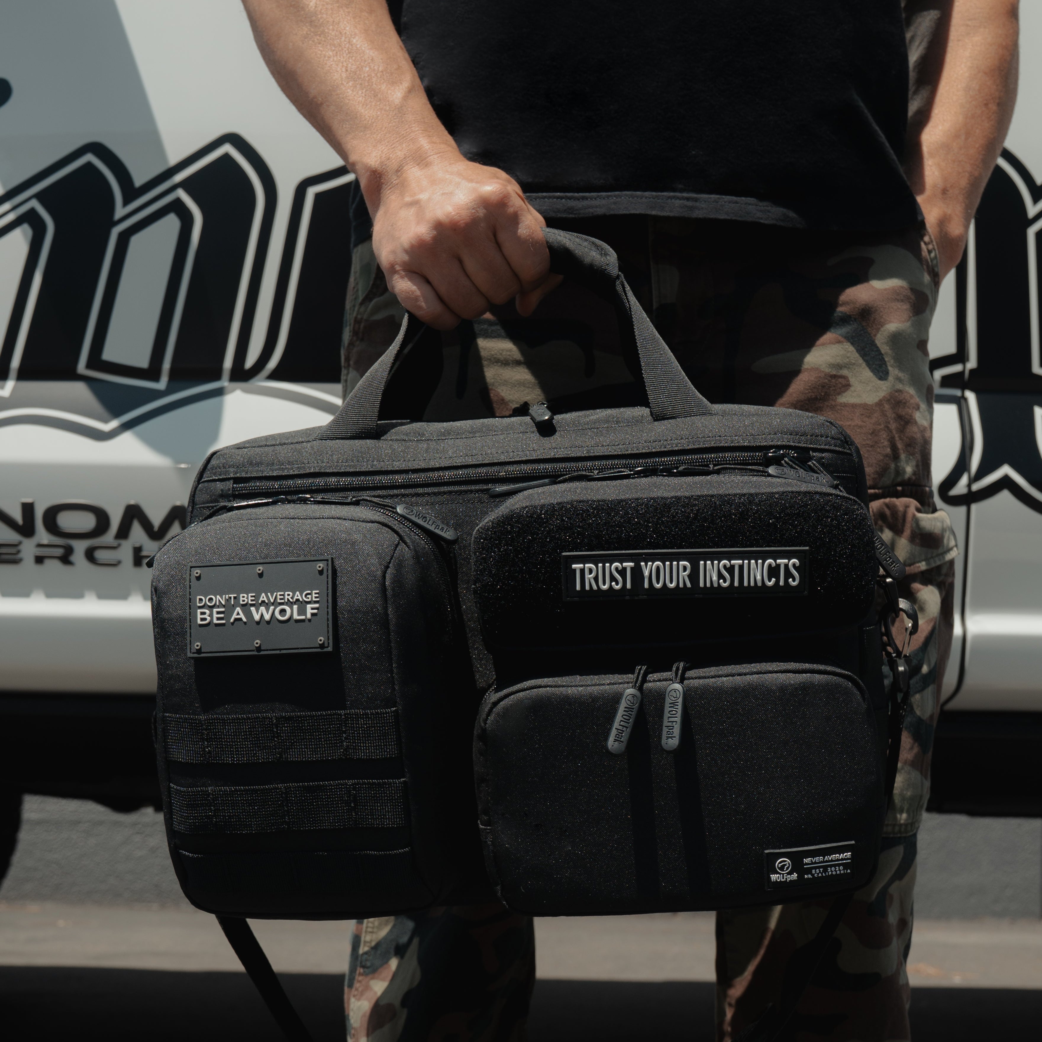 A person outdoors carrying a black tactical-style duffel bag with multiple front pockets, top handles, and visible branding patches. The bag has white text reading 'TRUST YOUR INSTINCTS' and a smaller patch that appears to say 'THE WOLF'.