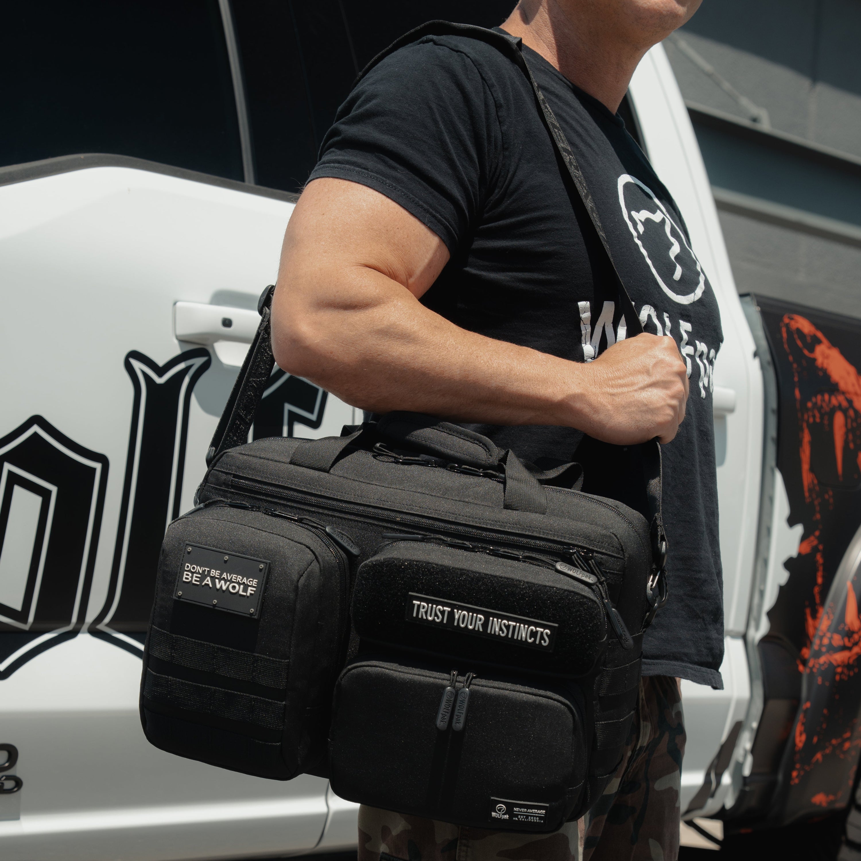 A man wearing a black t-shirt and camouflage pants carries a black tactical shoulder bag slung across his body, standing next to a white truck.
