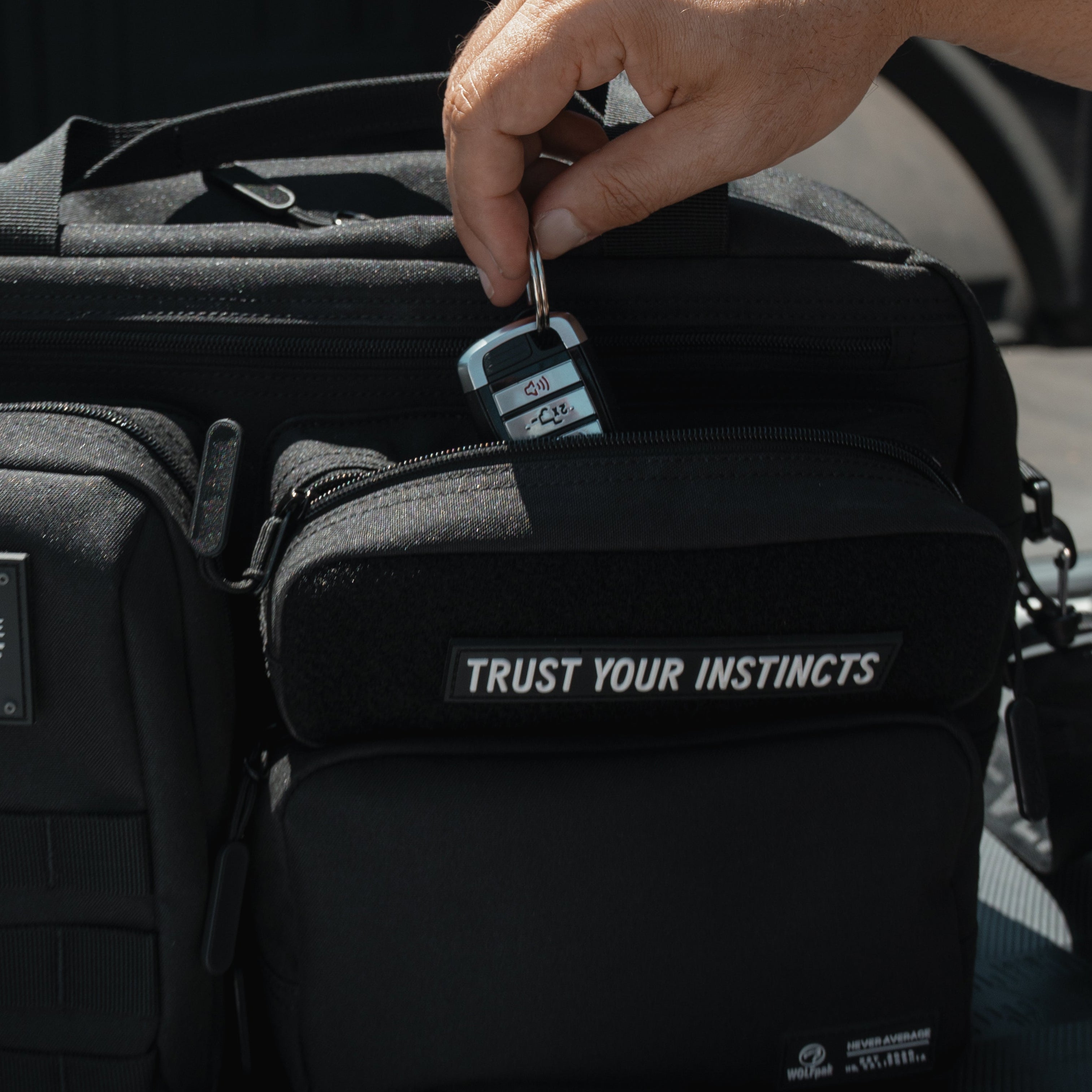 A black tactical-style bag with two front pockets sits in the bed of a truck. A hand is inserting a key into the lock on the front pocket.