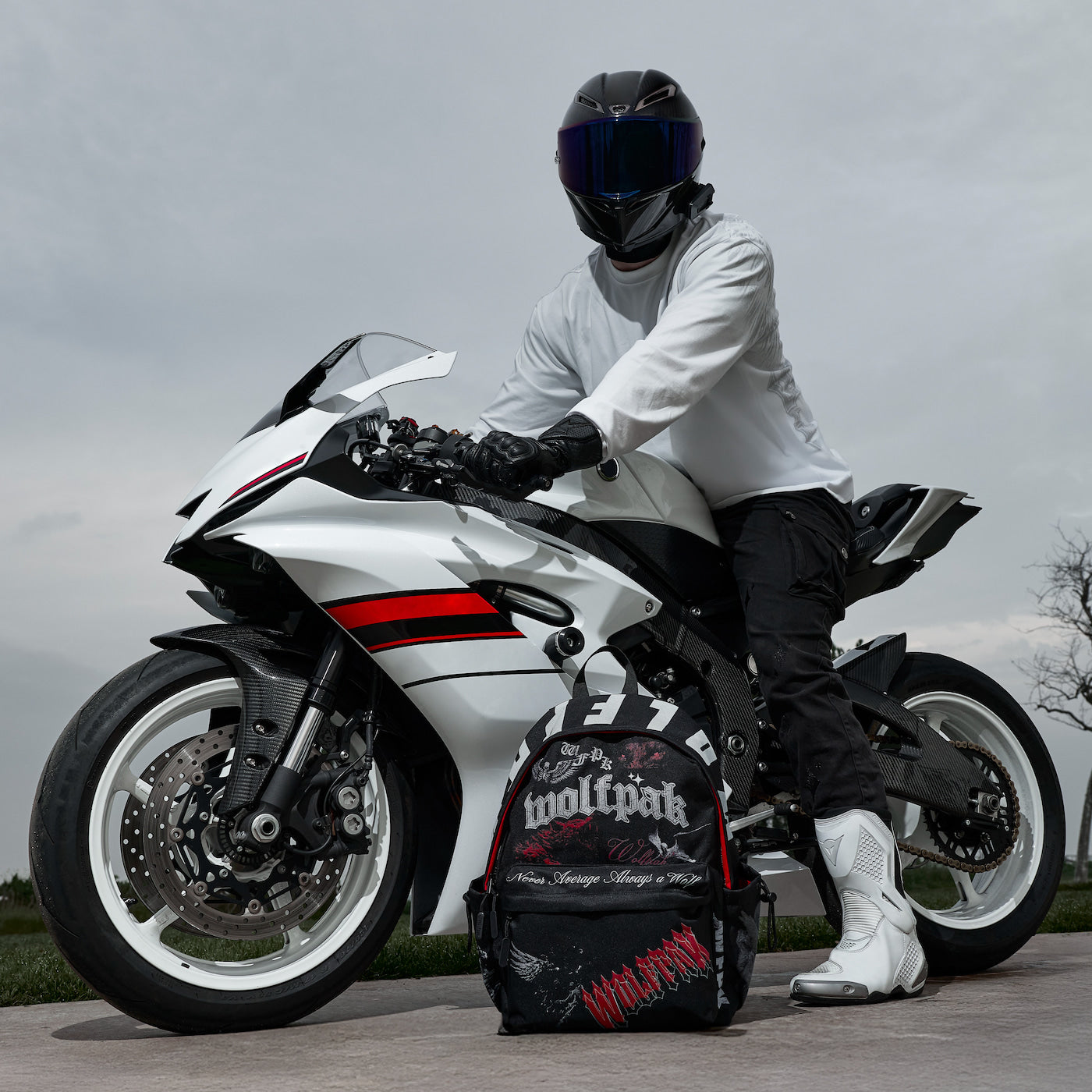 A person in a white long-sleeve shirt, black pants, gloves, and a black helmet sits on a white and black sport motorcycle outdoors. A black and red motorcycle bag with the brand name 'woolpak' rests on the ground in front of the bike.