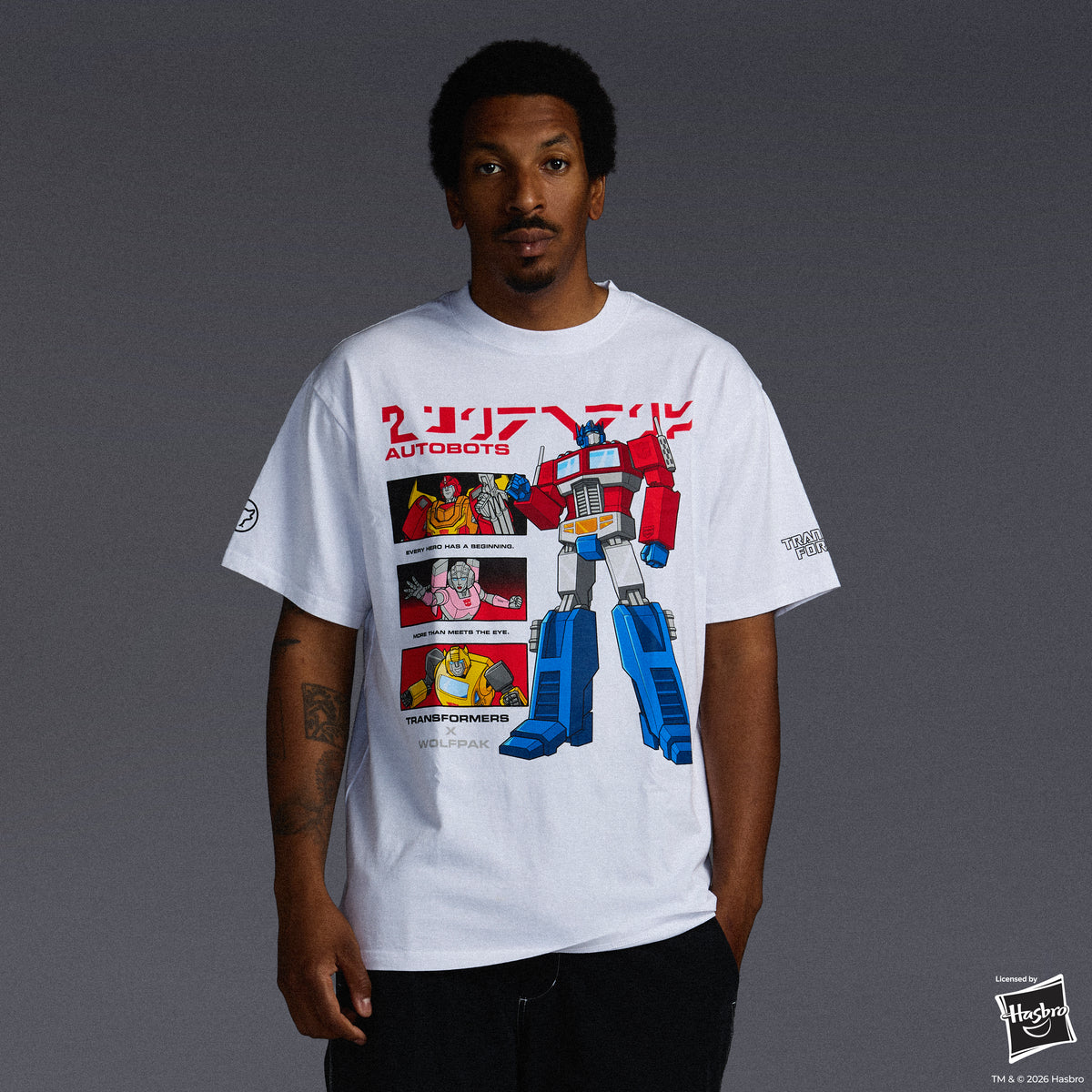 A man wearing a white graphic t-shirt featuring a red and blue Optimus Prime design with comic panels, photographed against a dark gray studio background.