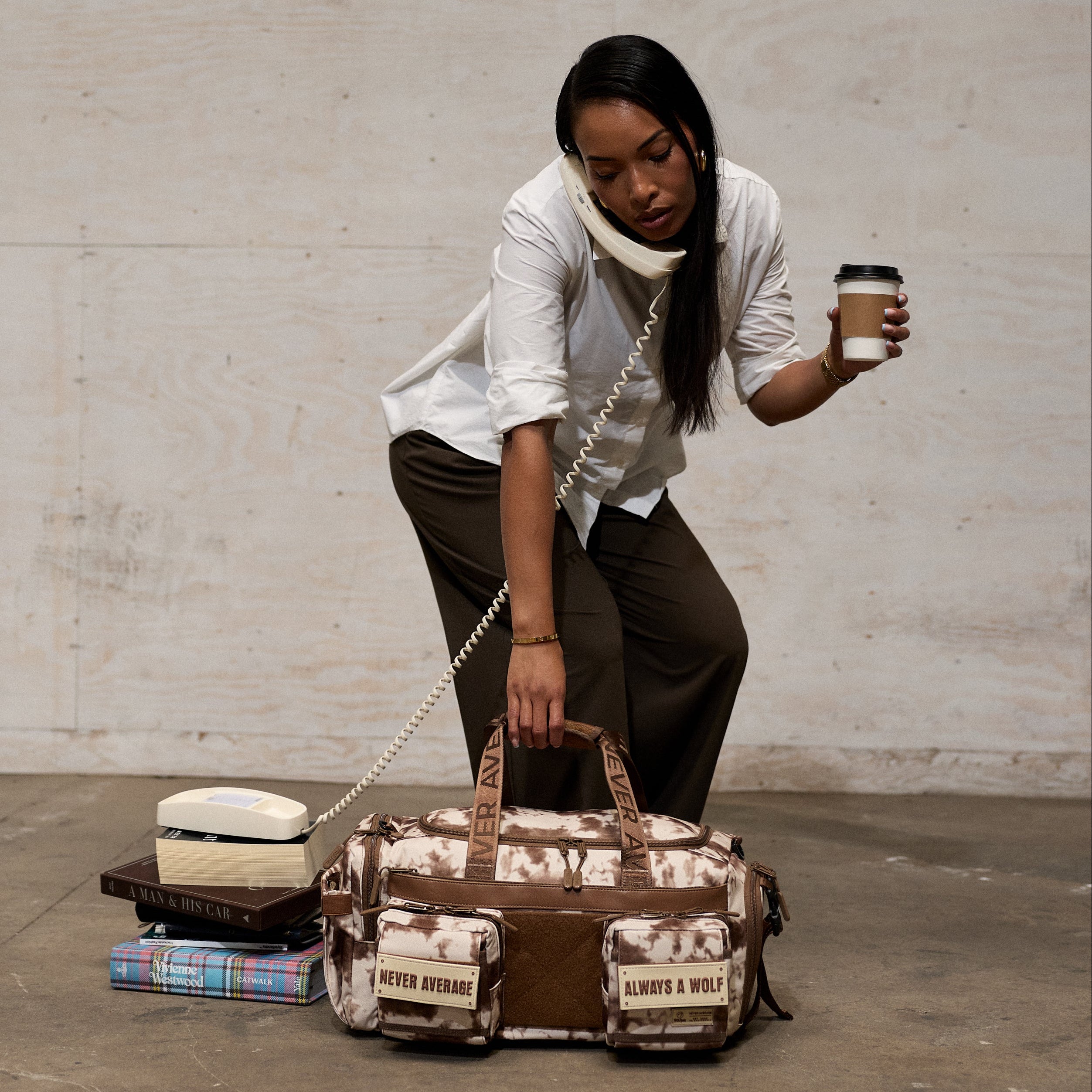 A woman in a white shirt and dark pants kneels beside a brown duffel bag with a beige and brown camouflage print, holding a coffee cup.