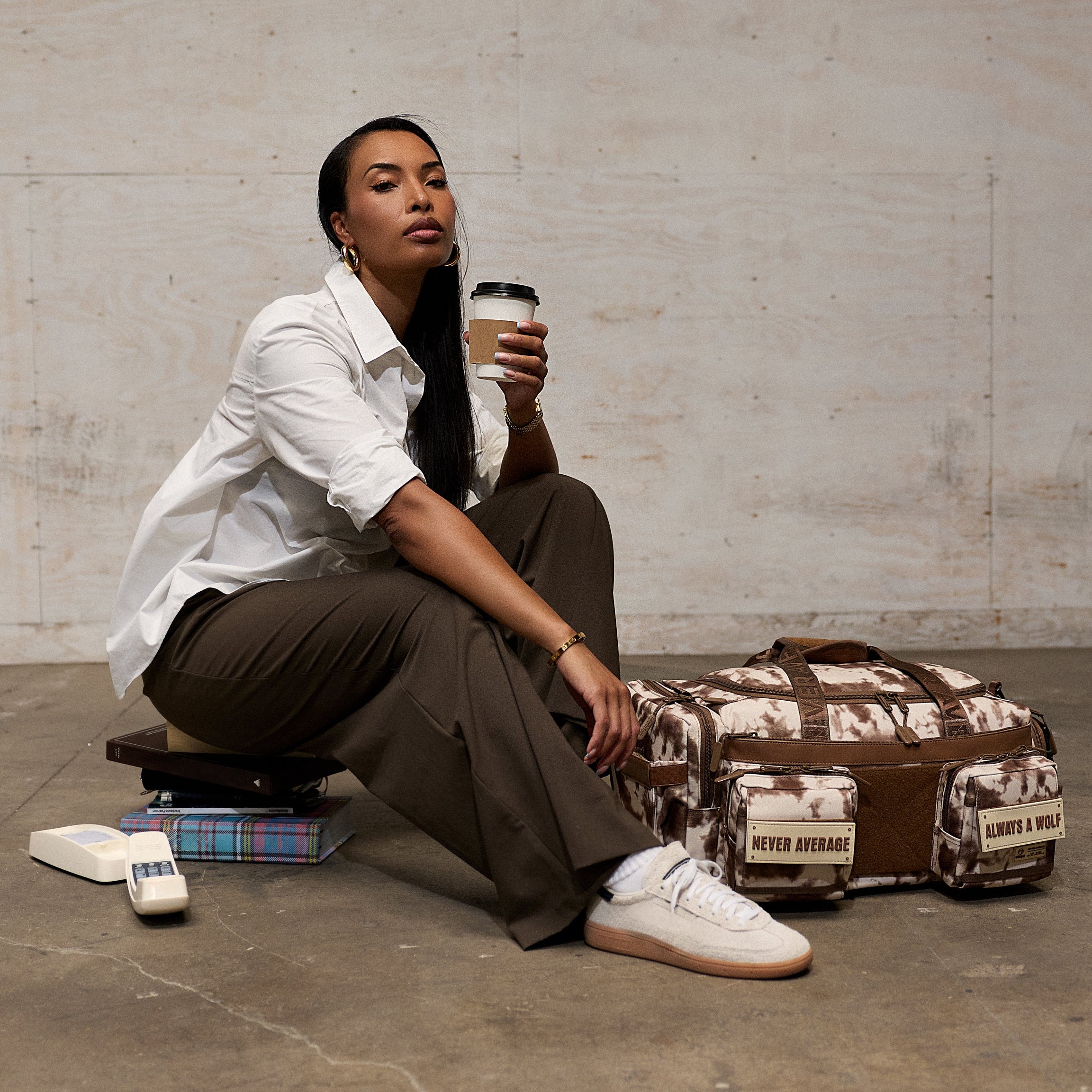 A woman sits on the floor holding a coffee cup next to a brown and beige patterned duffel bag with 'CITY & RUG' labels, in a minimalist concrete room.