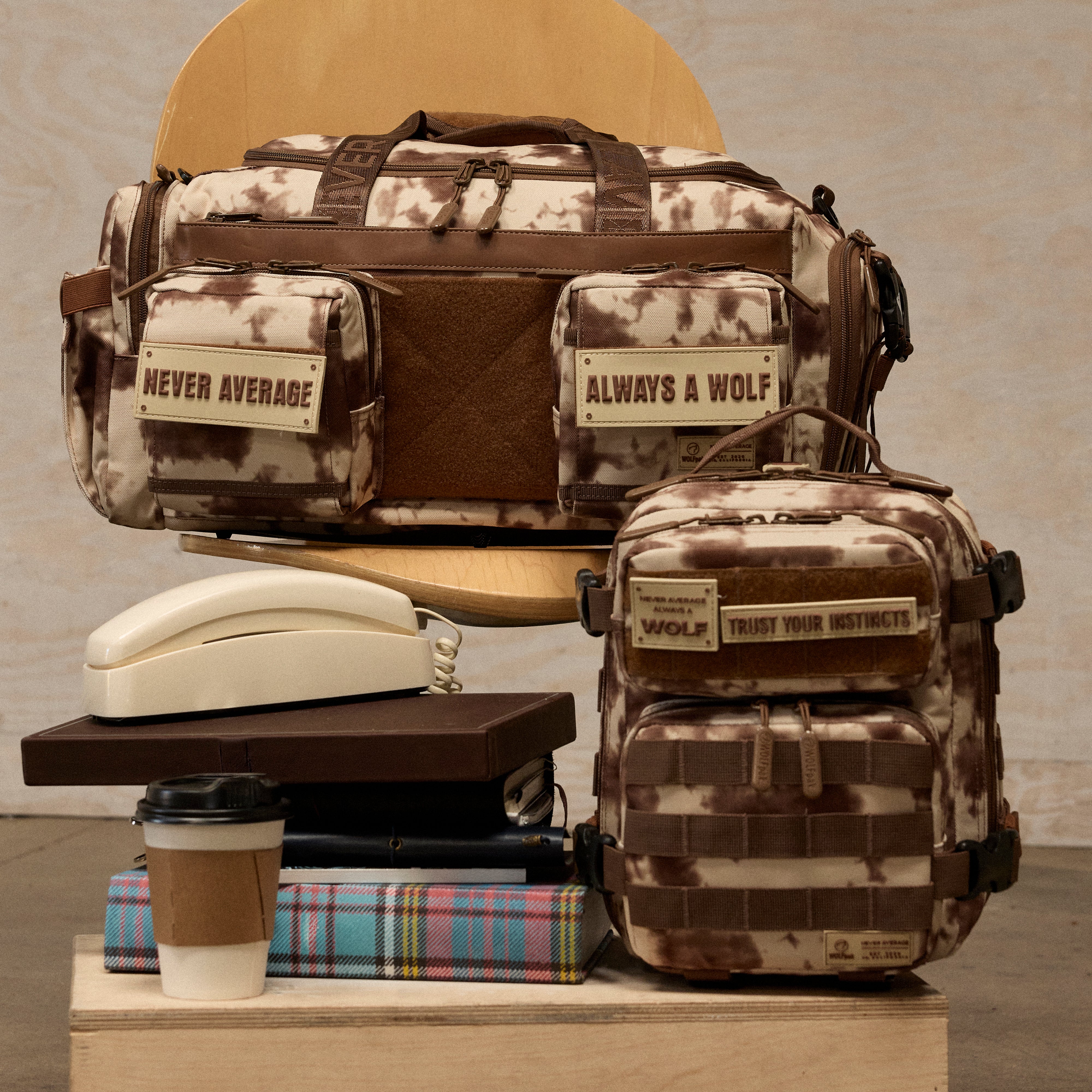 A camouflage duffel bag and matching backpack displayed together on stacked props in an indoor studio setup. The bags have brown leather-like trim and front patches with the phrases “NEVER AVERAGE” and “ALWAYS A WOLF,” plus a smaller patch reading “TRUST YOUR INSTRUCTORS.