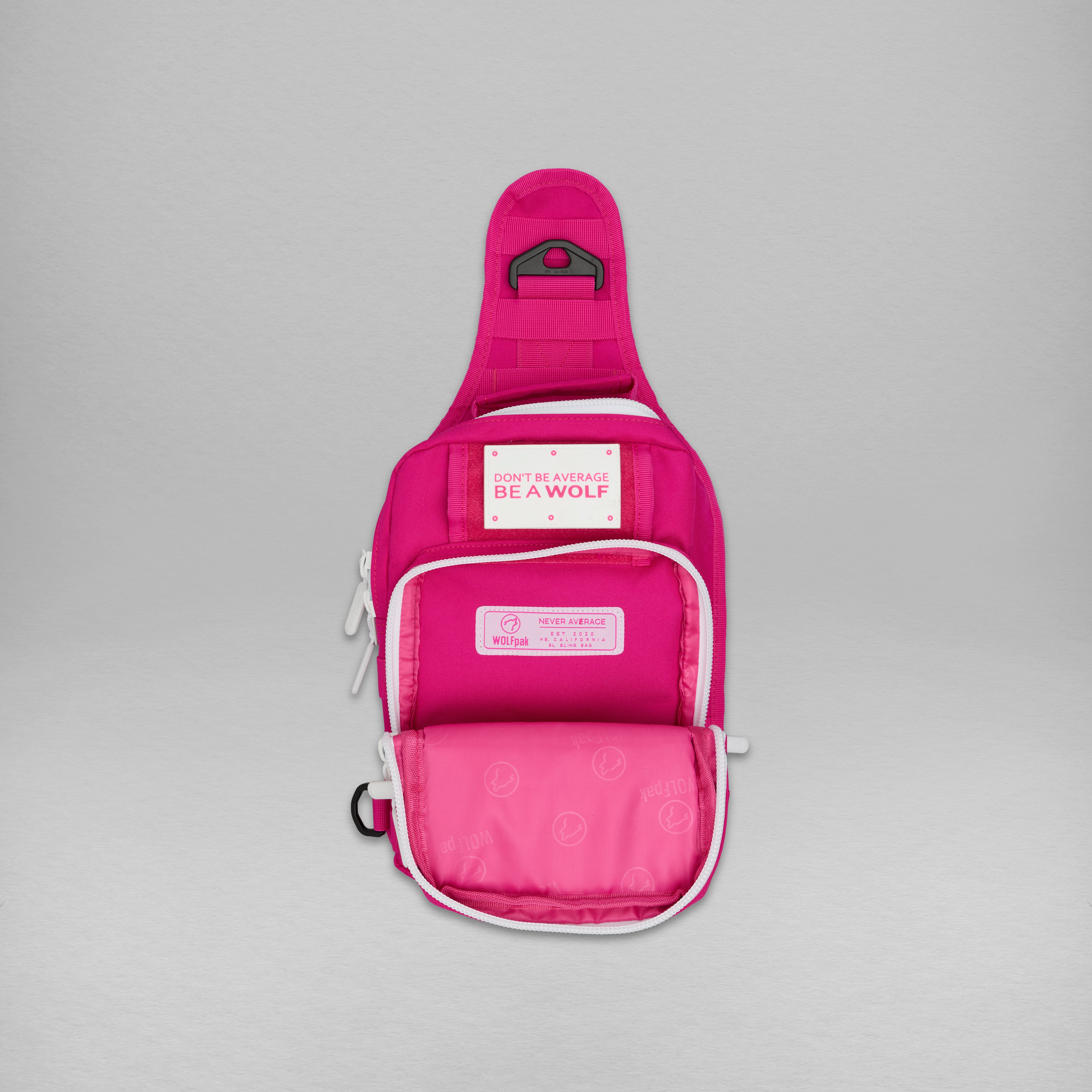 A bright pink sling backpack by Bella + Canvas, shown open against a neutral gray background. The bag features white piping, multiple zippered pockets, and a front patch reading 'BE A WOLF'.