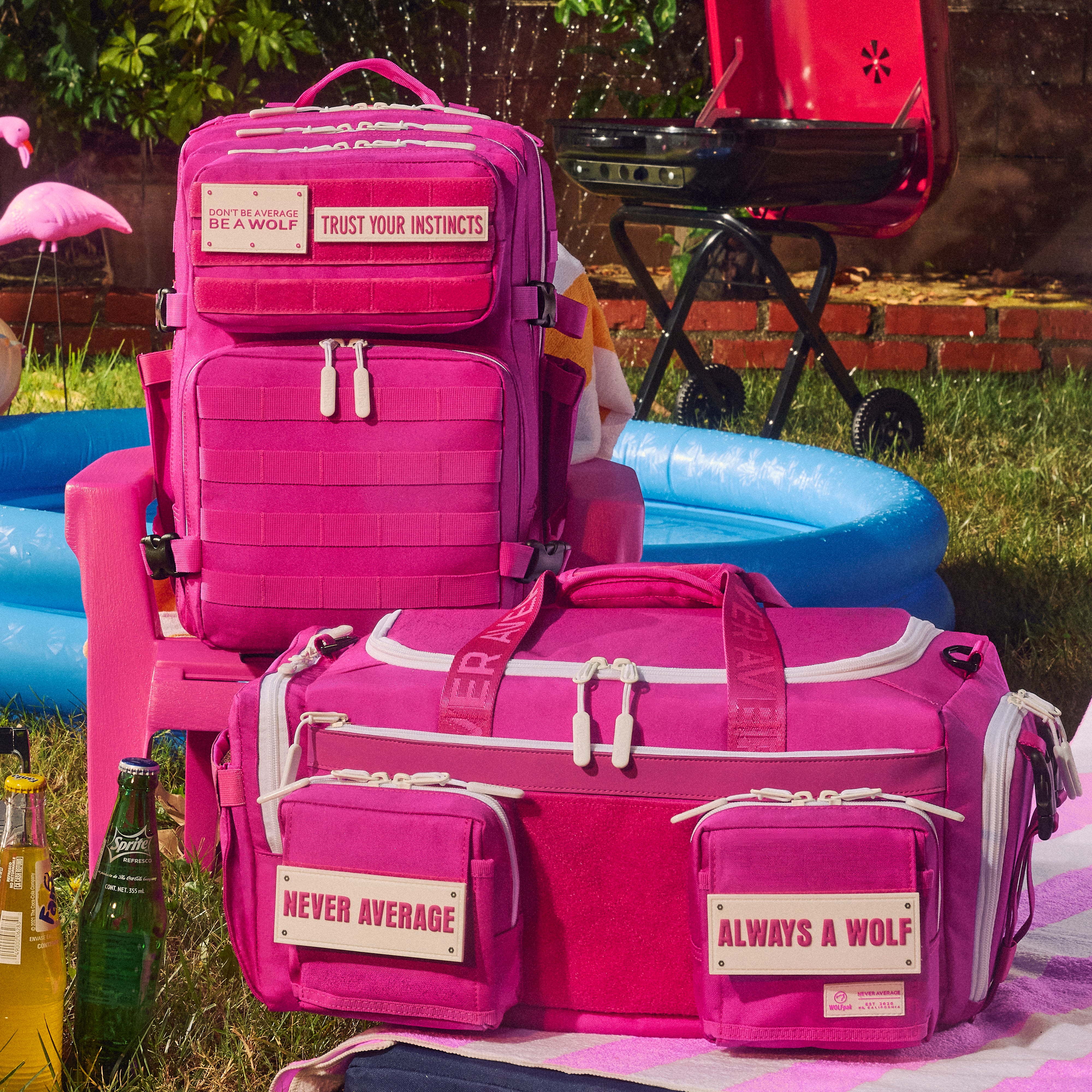 A bright pink backpack and matching duffel bag with white trim and slogan patches, styled outdoors on a lawn beside a pink chair, blue pool float, and barbecue grill.