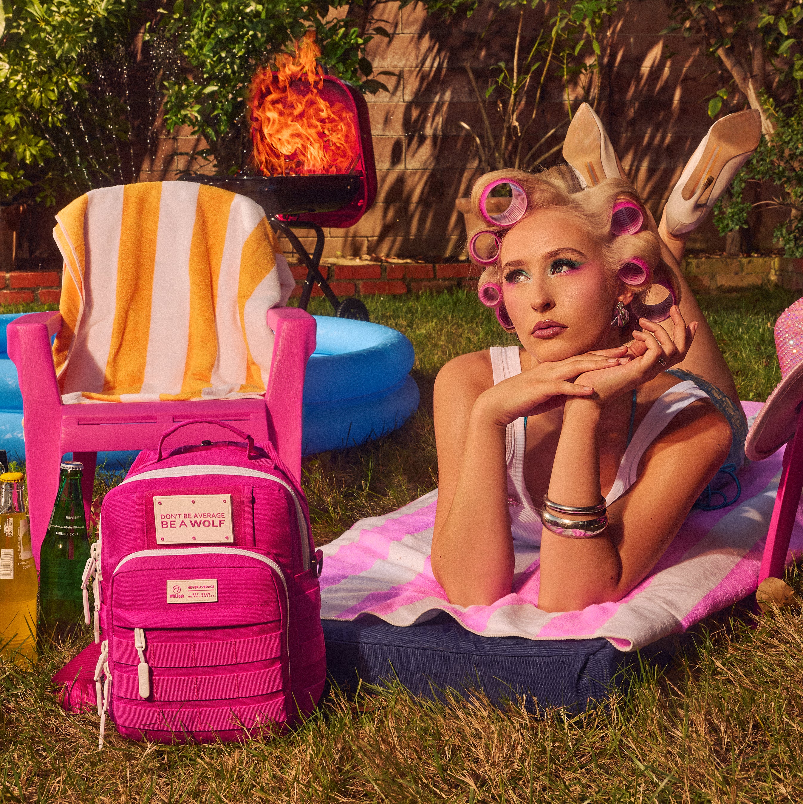 A woman reclines on a pink-and-white striped blanket in a backyard setting, wearing bunny ears and pink hair rollers. A bright pink backpack sits in the foreground on grass, with a pink chair, blue pool float, and a flaming grill in the background.