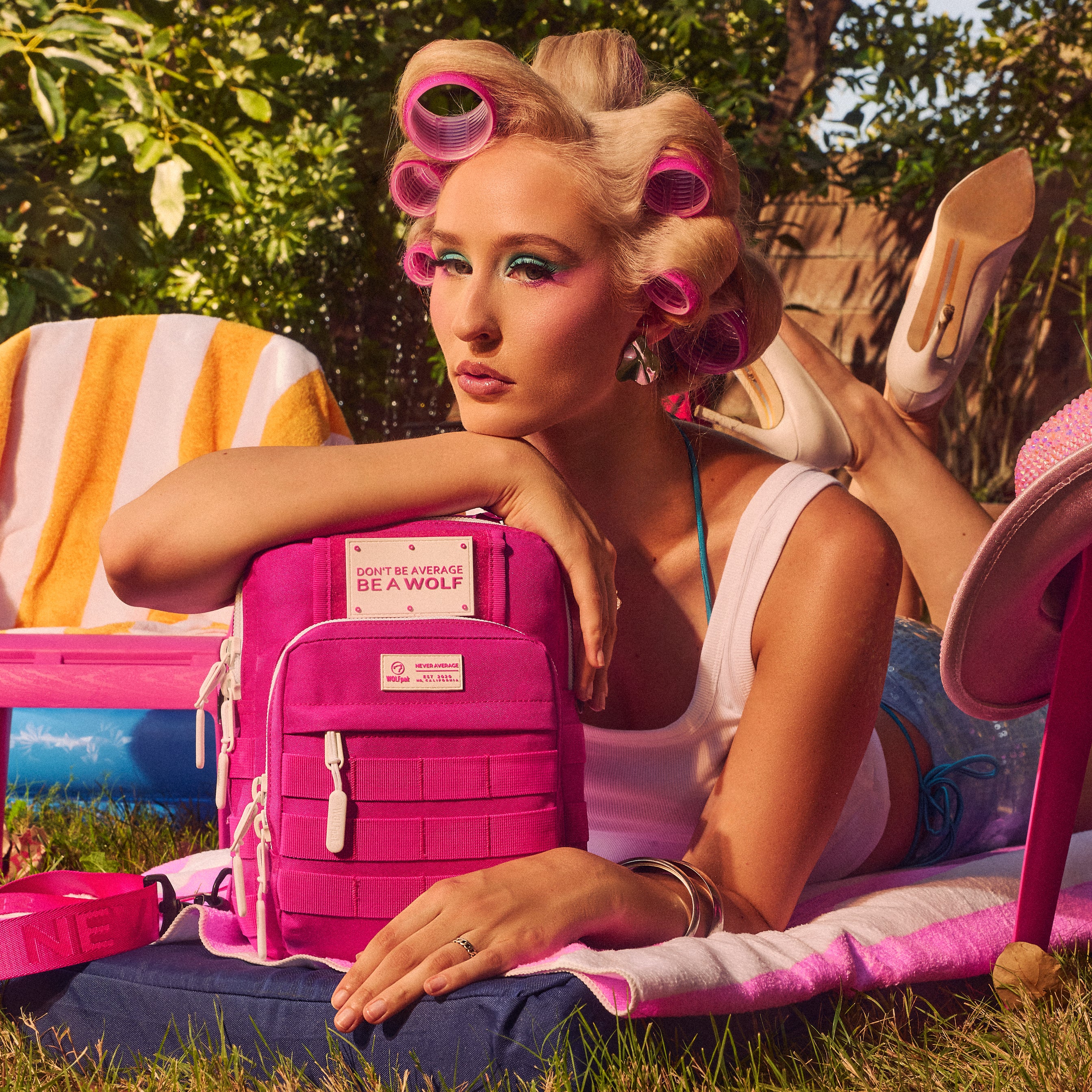 A woman reclining outdoors on a blanket with pink hair rollers, a pink backpack, and pink accessories in a sunny garden setting. The backpack has a visible front patch reading 'GOT THE ENERGY BE A WOLF' and a small white logo label.