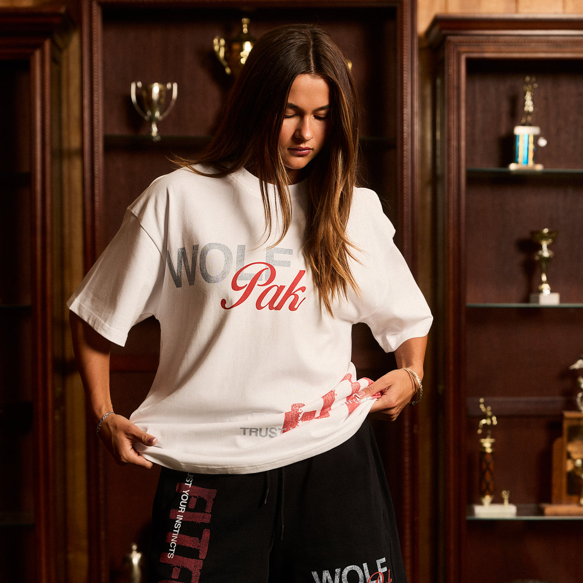 A woman wearing an oversized white graphic T-shirt with 'WOLFpak' printed across the chest in gray and red, paired with black sweatpants, standing in front of a dark wood trophy cabinet.