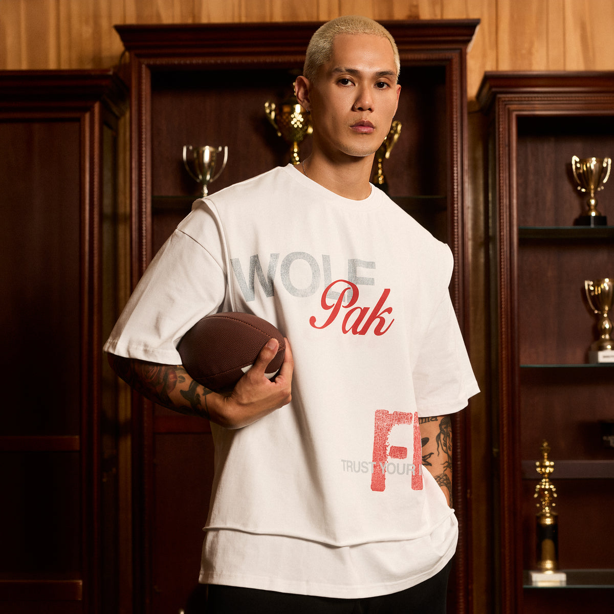 A man wearing a white oversized T-shirt with 'WOLFPak' printed in gray and red, holding a football in front of a dark wood trophy cabinet.
