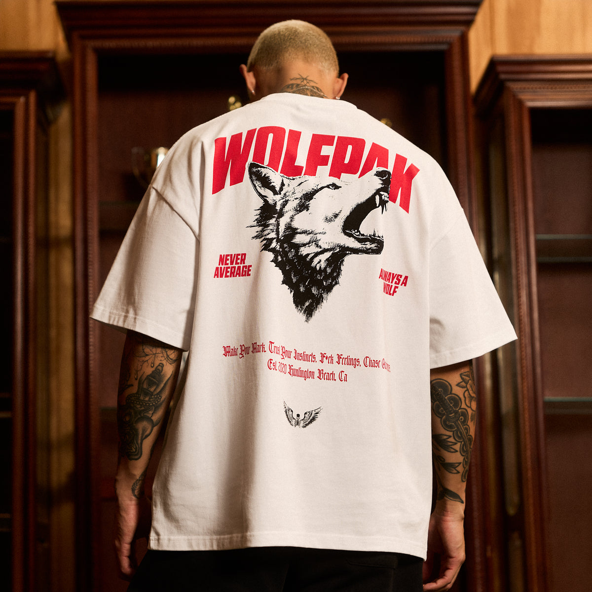 A man viewed from behind wearing an oversized white t-shirt with a large red and black wolf graphic and 'WOLFPAK' text on the back, standing in front of dark wooden cabinets.