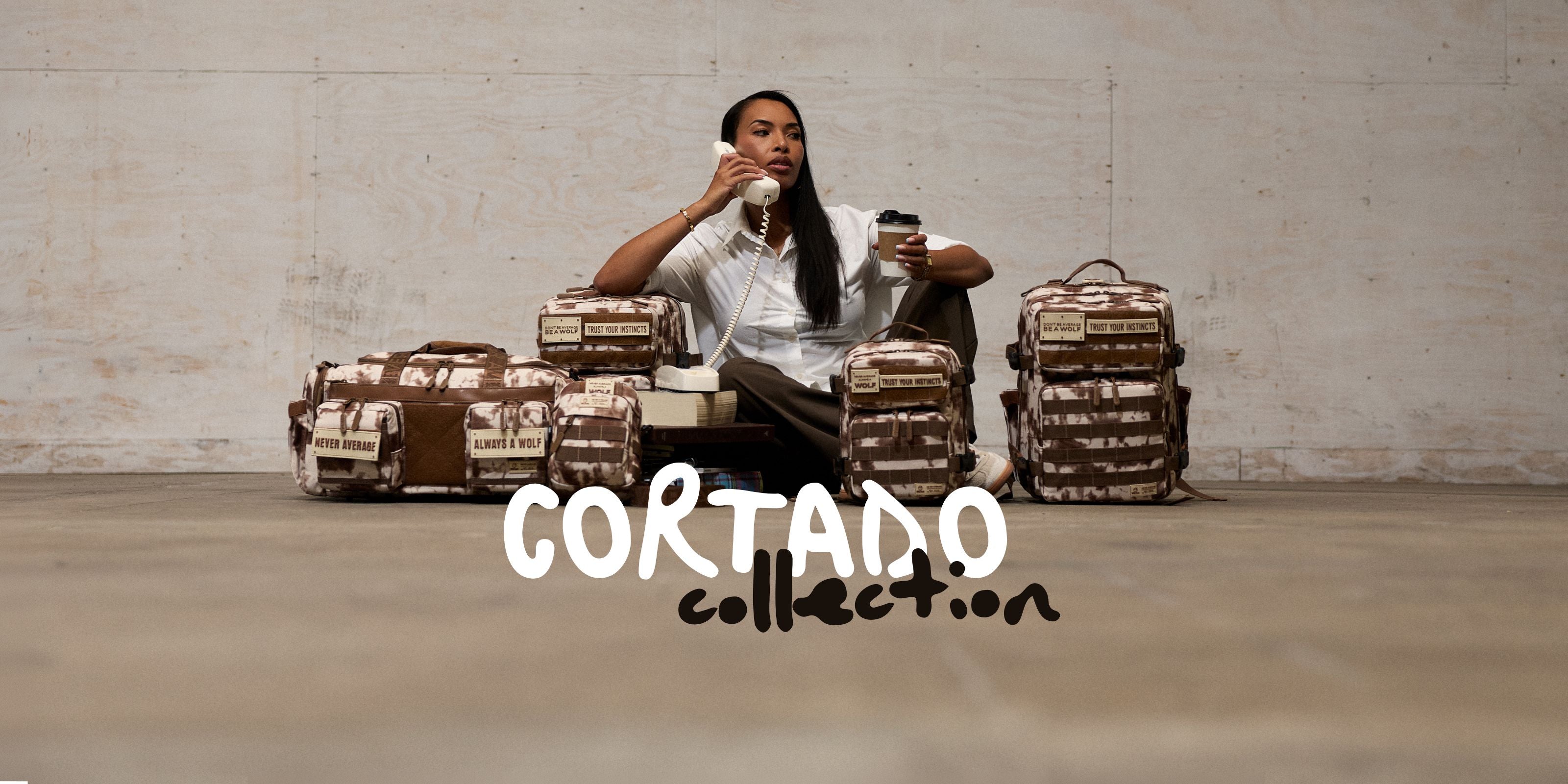 A person sits on the floor in front of a concrete wall, surrounded by multiple Cortado Collection backpacks and bags in a brown and beige camouflage pattern. The person holds a coffee cup and a phone, with the text 'CORTADO collection' overlaid at the bottom.