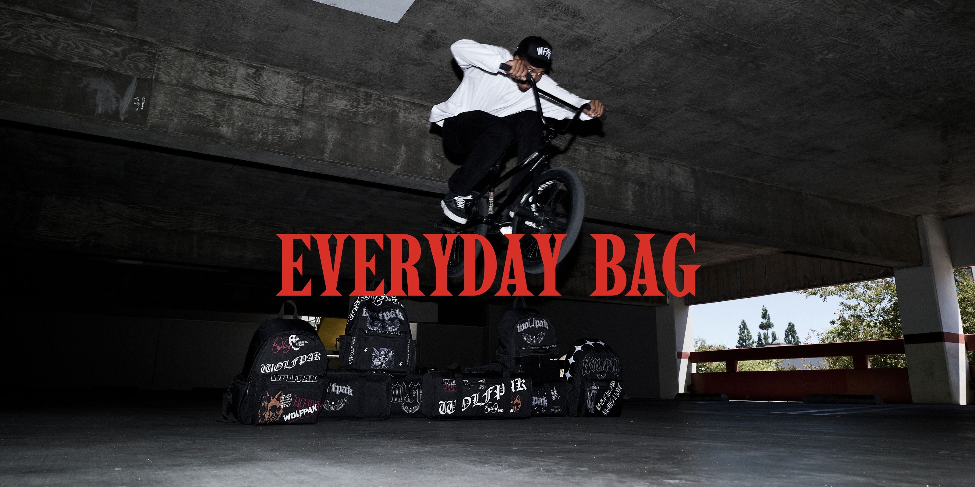 A BMX rider in a white shirt and black pants jumps over a bike under a concrete bridge, with several black Everyday Bag backpacks displayed below. Large red text reads 'EVERYDAY BAG' across the center.