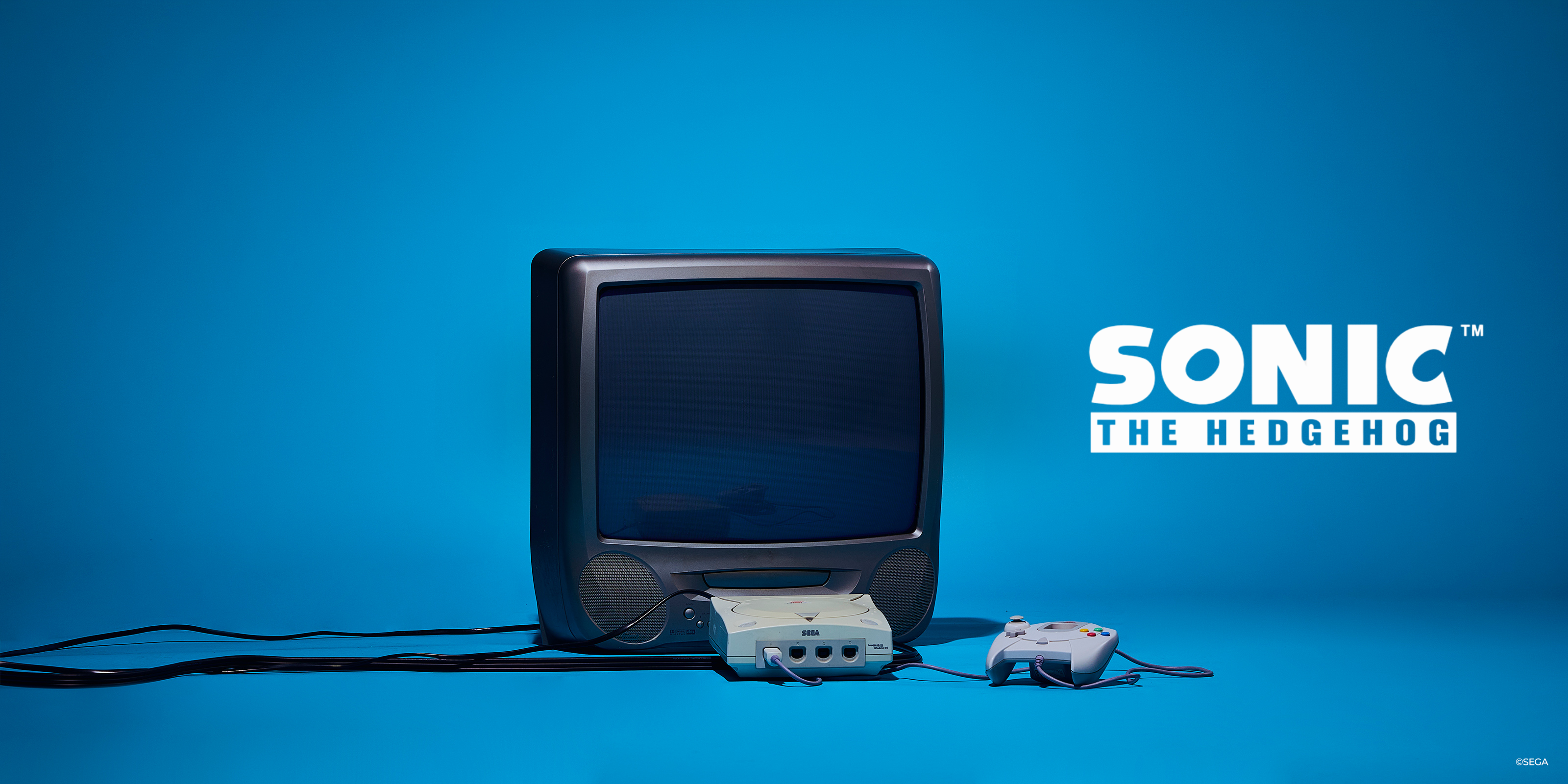 A retro-style television console setup with a gray controller on a bright blue background, alongside the Sonic the Hedgehog logo. The console resembles a vintage CRT TV with a built-in controller dock and a matching controller placed to the right.