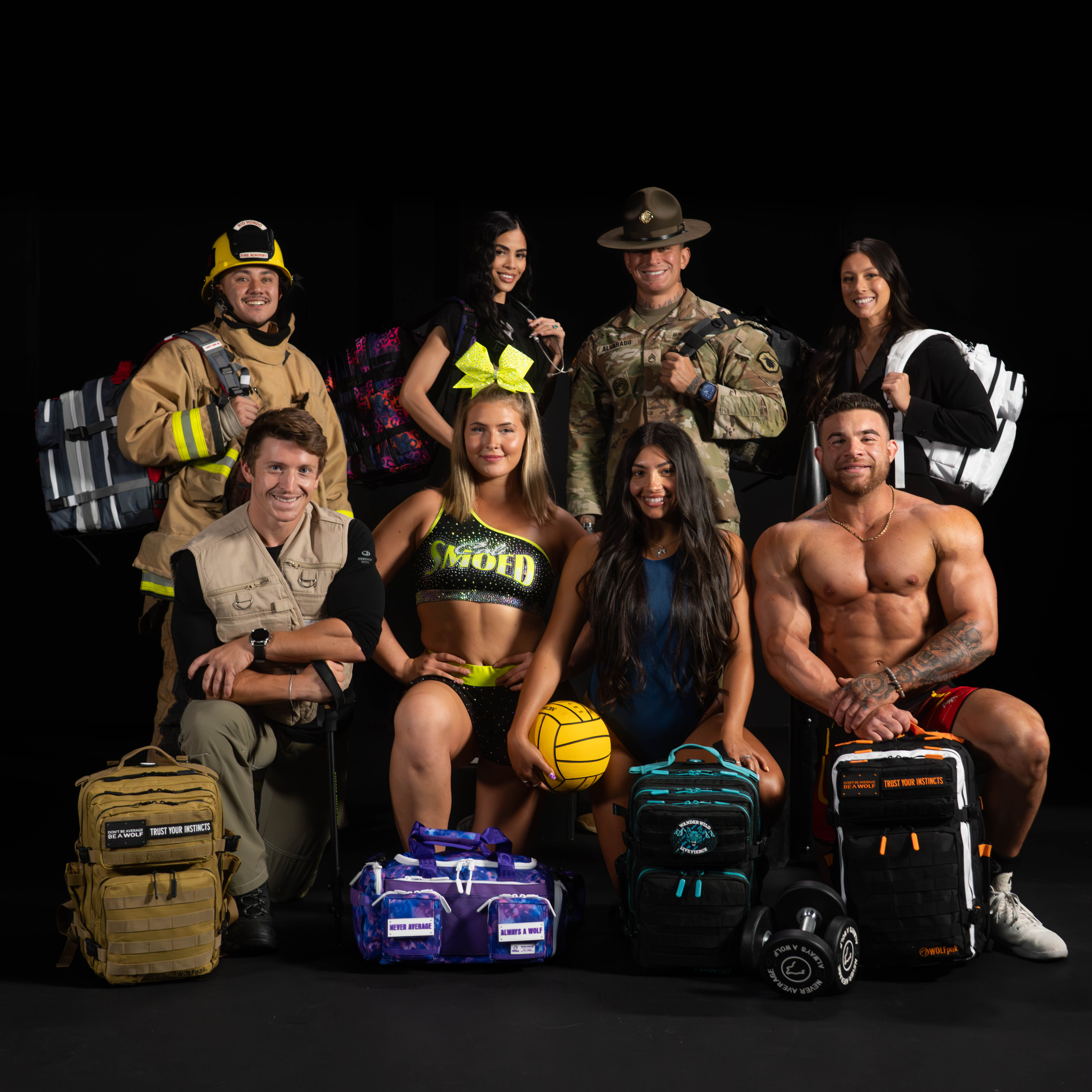 A group of eight people posing with various backpacks and duffel bags against a solid black background. The group includes individuals in firefighter, military, and athletic attire, along with several bags in tan, purple, teal, and black.